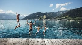 Čtyři lidé skáčou z mola do jezera v Zell am See-Kaprun, v pozadí hory a město.