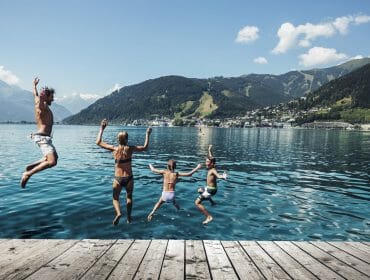 Čtyři lidé skáčou z mola do jezera v Zell am See-Kaprun, v pozadí hory a město.