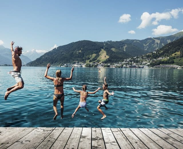 Čtyři lidé skáčou z mola do jezera v Zell am See-Kaprun, v pozadí hory a město.