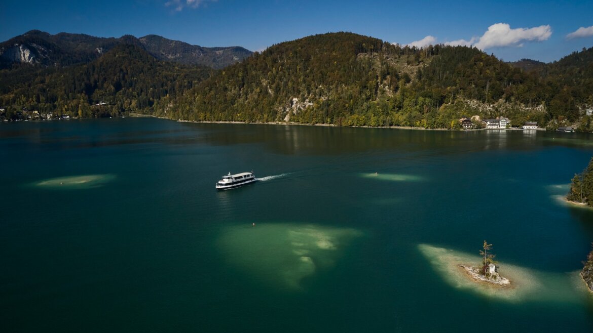 Loď pluje po Wolfgangsee, modrém jezeře obklopeném zalesněnými kopci a ostrůvky pod jasnou oblohou.
