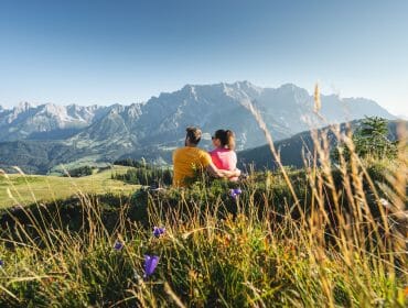Pár sedí na rozkvetlé horské louce a kochá se pohledem na majestátní štíty Hochkönigu