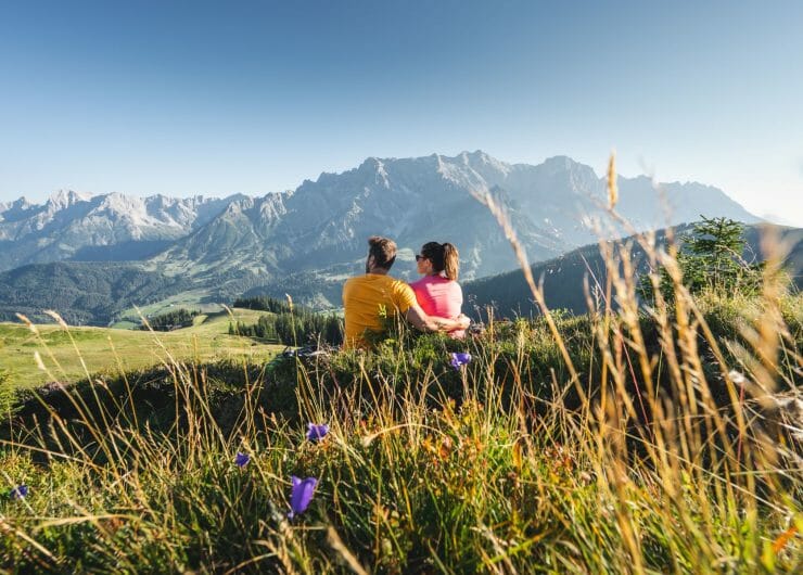 Pár sedí na rozkvetlé horské louce a kochá se pohledem na majestátní štíty Hochkönigu