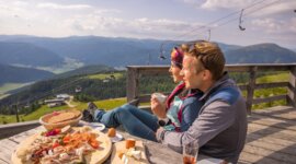 Dvojice turistů si užívá pauzu na terase horské chaty v Salcburském Lungau s lahodnou svačinou z regionálních specialit. V povzdálí se vznášejí letní lanovky.