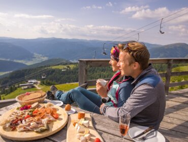 Dvojice turistů si užívá pauzu na terase horské chaty v Salcburském Lungau s lahodnou svačinou z regionálních specialit. V povzdálí se vznášejí letní lanovky.