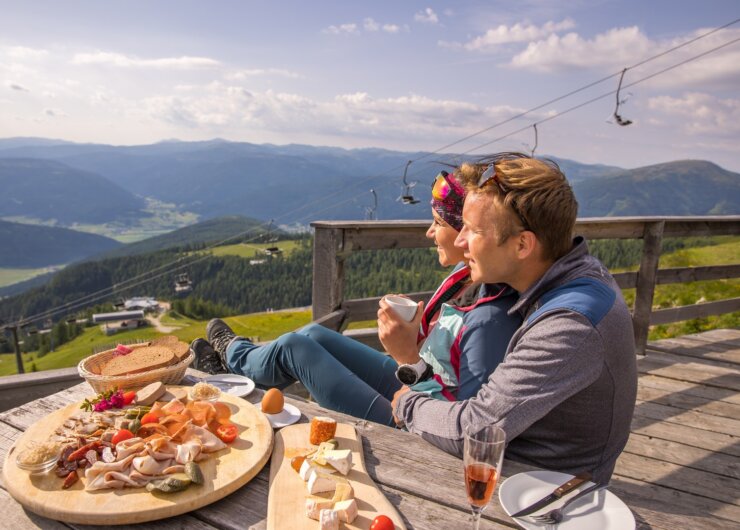 Dvojice turistů si užívá pauzu na terase horské chaty v Salcburském Lungau s lahodnou svačinou z regionálních specialit. V povzdálí se vznášejí letní lanovky.