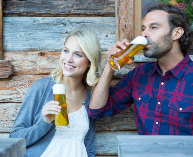 Usměvavá žena a muž si společně užívají pivo před rustikálním dřevěným stavením.