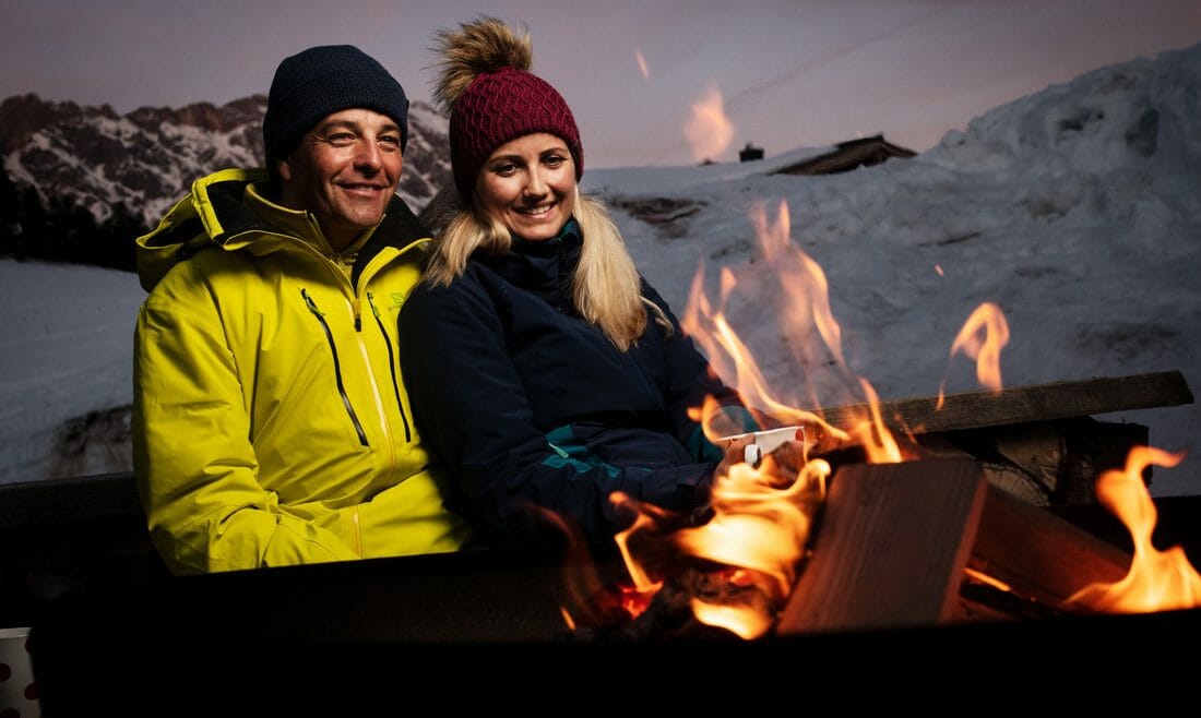 Pár sedí večer venku u plápolajícího ohně v zimní rekreační oblasti Hochkönig