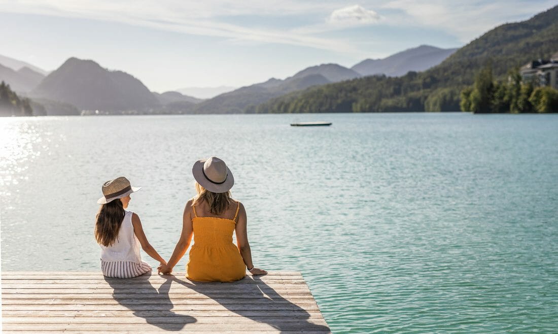 Dva lidé v kloboucích sedí na dřevěném molu u jezera, na slunném pozadí jsou hory a vodní sporty.