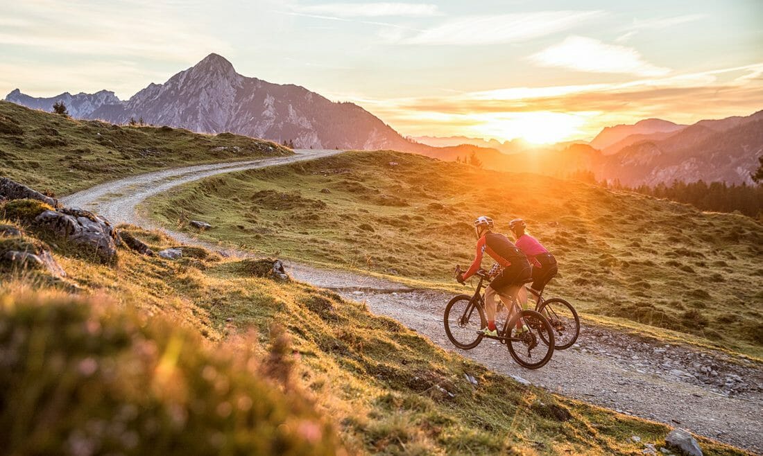 Cyklisté jedou po šotolinové cestě mezi zelenými pastvinami při západu slunce v regionu Dachstein-West