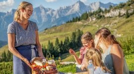 Hostitelka přináší ženě s dětmi na stůl prkénko se sýry, uzeninami, pomazánkami a máslem