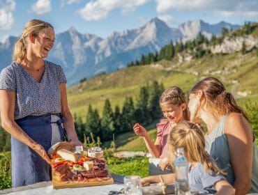 Hostitelka přináší ženě s dětmi na stůl prkénko se sýry, uzeninami, pomazánkami a máslem