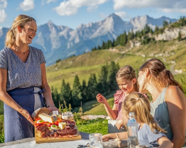 Bei der Jagglhütte Hostitelka přináší ženě s dětmi na stůl prkénko se sýry, uzeninami, pomazánkami a máslem