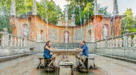 Dva lidé sedí u kamenného stolu u hravých fontán v zahradě plné soch nedaleko historických Hradů.