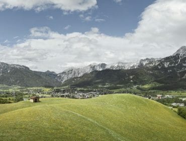 Zelené kopce s malou chatou, městečkem a zasněženými horami pod částečně zataženou oblohou.