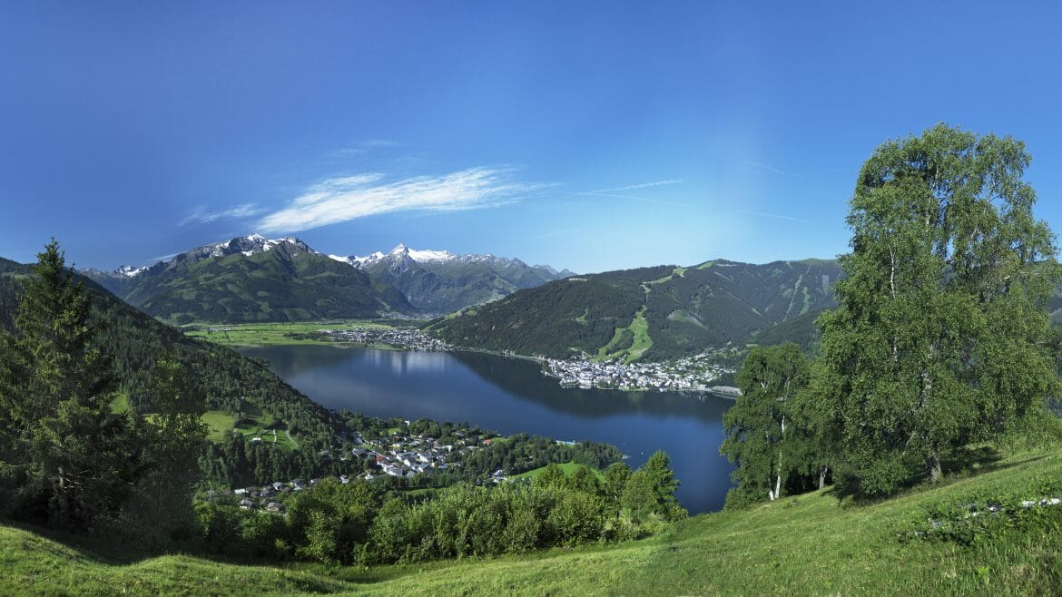 Klidné jezero v Zell am See-Kaprun, rámované zelenými kopci a zasněženými horami pod jasně modrou oblohou. (Zvětšený pohled)