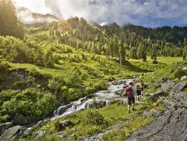 Turisté kráčejí podél kamenitého potoka ve svěžích zelených horách Saalbach Hinterglemm pod částečně zataženou oblohou.