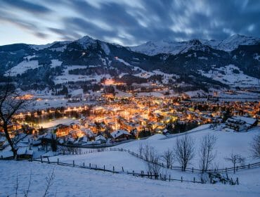 Zasněžená vesnice Gastein v údolí za soumraku, zářící světla a hory v pozadí.