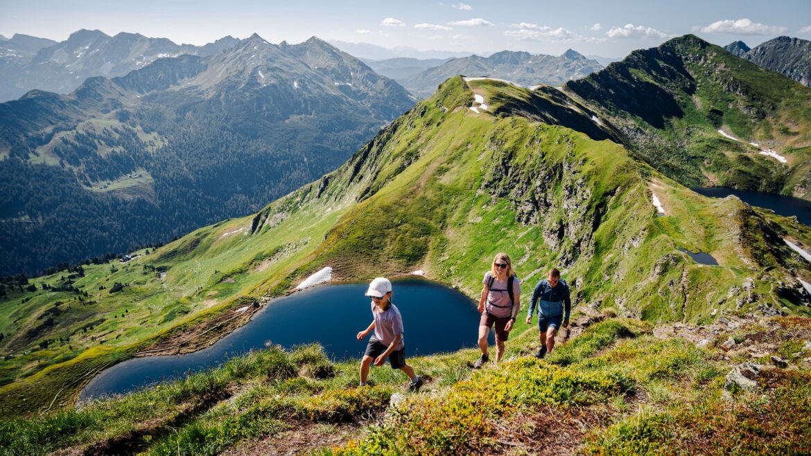 Rodina si užívá jednu z mnoha turistických tras v salcburském Lungau. V pozadí je vidět okolní horská krajina a horské jezero „Twenger Almsee“.