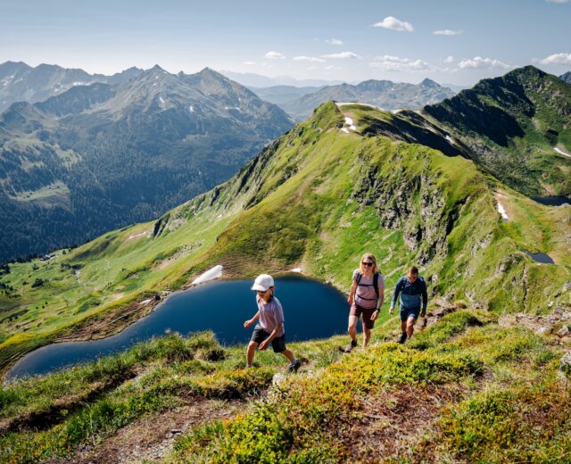 Rodina si užívá jednu z mnoha turistických tras v salcburském Lungau. V pozadí je vidět okolní horská krajina a horské jezero „Twenger Almsee“.