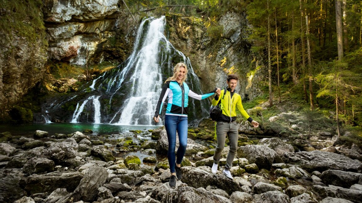 Dva lidé na túře na skalách u vodopádu v Tennengau Salcbursko, drží se za ruce a usmívají se.