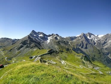 Klikatá horská silnice se zelenými kopci, červeným autem a zasněženými vrcholky pod jasně modrou oblohou.