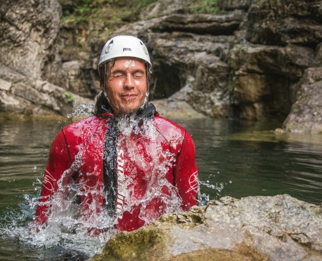 Osoba v červeném neoprenu a bílé helmě se vynořuje z vody ve skalnatém prostředí adrenalinového sportu na Salcbursku.