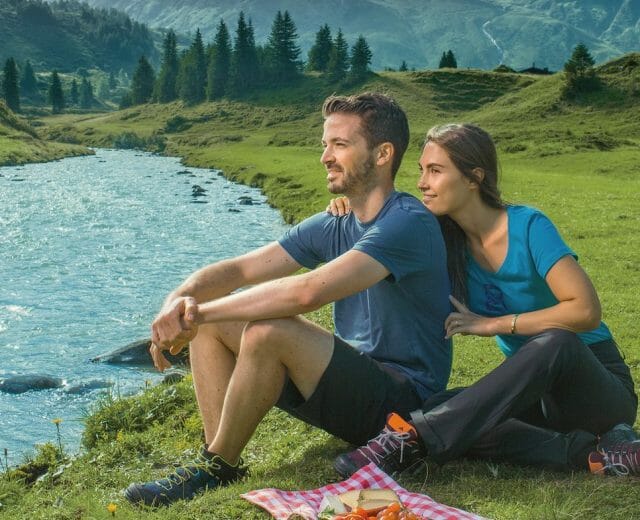 Pár piknikuje u řeky s horami za zády, idylická scéna z Oficiální webové stránky Salcbursko.