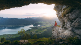Člověk sedí na skalním výběžku nad salcburským údolím, jezerem a horami při západu slunce