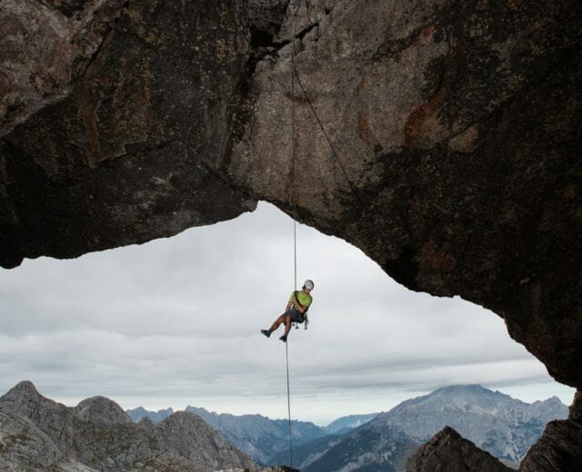Horolezec visí na laně pod skalním obloukem, v pozadí jsou hory a zatažená obloha.