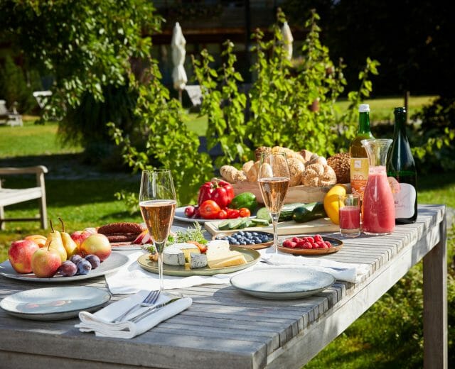 Venkovní dřevěný stůl s ovocem, sýrem, chlebem, nápoji a sklenicemi v zahradním prostředí.
