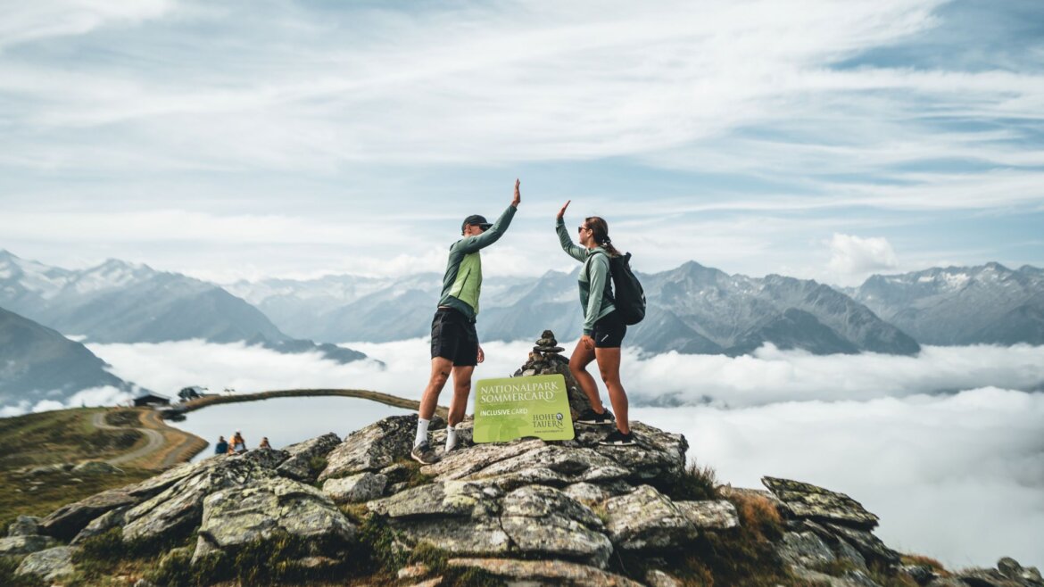 Dva turisté si plácají na skalnatém vrcholu ve Wildkogel Areně Neukirchen Bramberg, za nimi zasněžené hory.