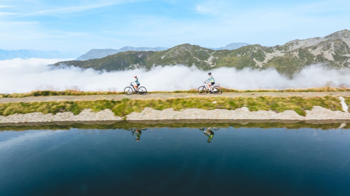 Dva lidé jedou na kolech po horské stezce vedle klidného jezera ve Wildkogel Arena Neukirchen Bramberg. (Zvětšený pohled)