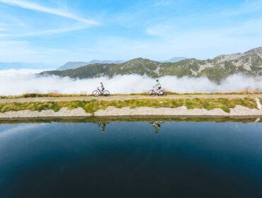 Dva lidé jedou na kolech po horské stezce vedle klidného jezera ve Wildkogel Arena Neukirchen Bramberg.