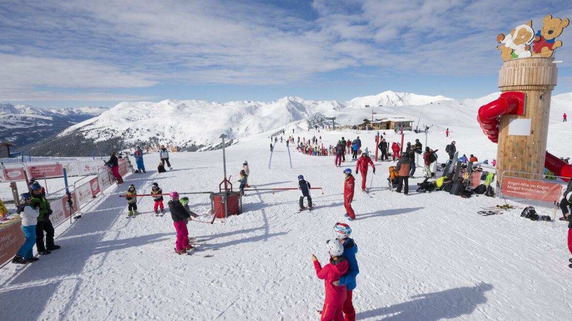 Lyžaři a děti si hrají na zasněžené sjezdovce Wildkogel Arena Neukirchen Bramberg u dětského hřiště pod modrou oblohou.