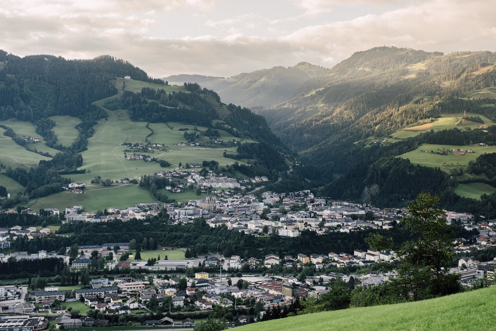 Stadtbild von St. Johann im Sommer Městečko St. Johann v zeleném horském údolí pod částečně zataženou oblohou v Salcbursku.