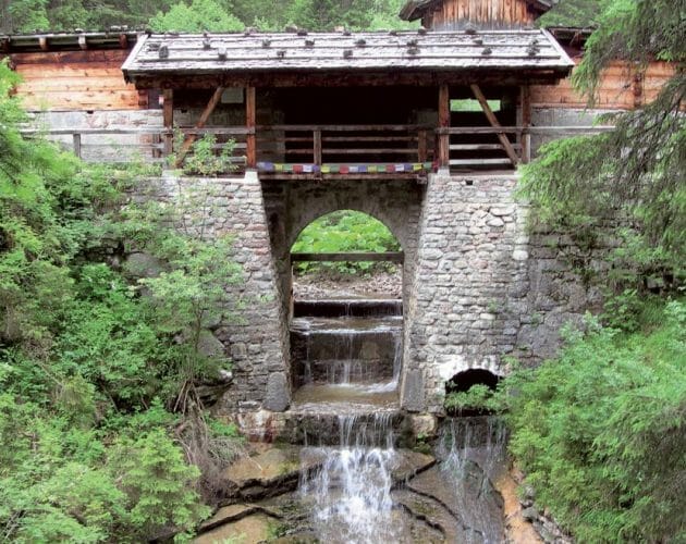 Rustikální dřevěný most s kamennou podezdívkou překračuje potok s malými vodopády ve svěže zeleném lese.