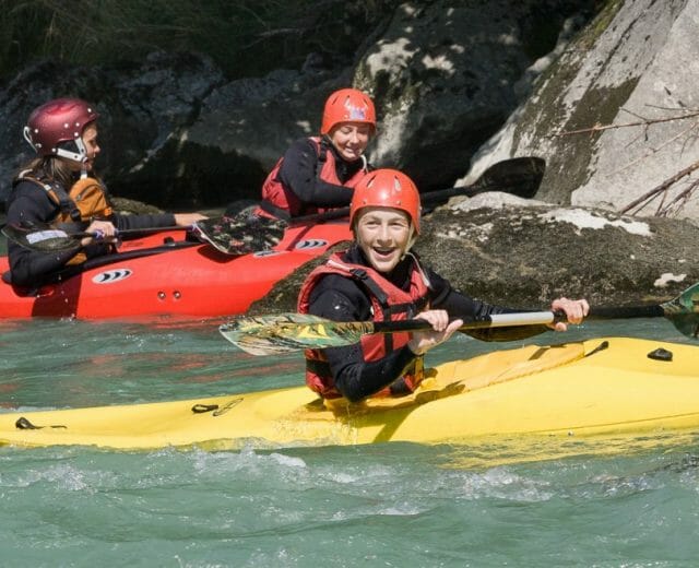 Tři lidé v barevných kajacích pádlují po řece, všichni mají helmy a záchranné vesty a usmívají se.