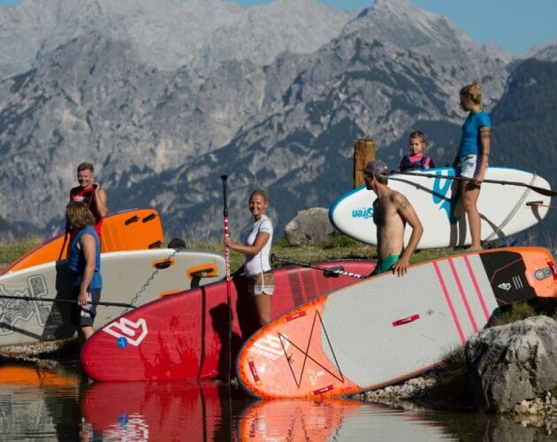 Čtyři lidé s paddleboardy stojí u horského jezera a usmívají se za slunečného dne.