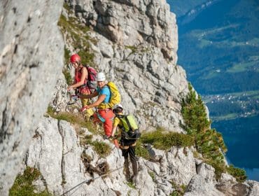 Tři horolezci s helmami a výstrojí stoupají po horské stezce v Solné komoře nad malebným údolím pod nimi.