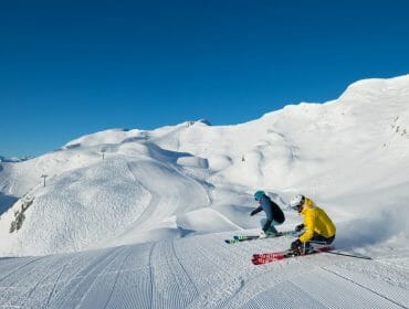 Dva lyžaři ve světlé výstroji se připravují na sjezd po čerstvě upraveném zasněženém svahu pod jasnou modrou oblohou.