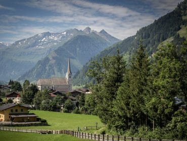 Vesnice s kostelní věží uprostřed zelených polí a alpských hor pod částečně zataženou oblohou.