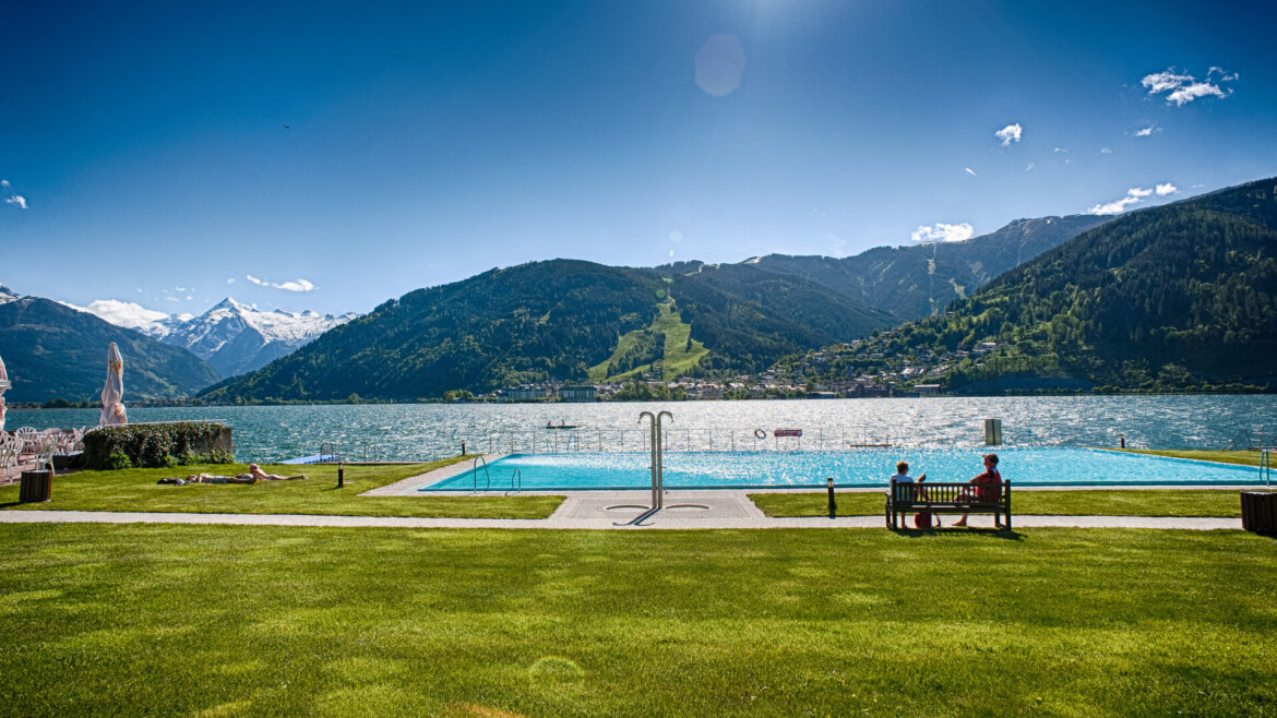 Venkovní bazén u jezera v Zell am See-Kaprun léto, hory a lidé odpočívající pod slunečnou modrou oblohou. (Zvětšený pohled)