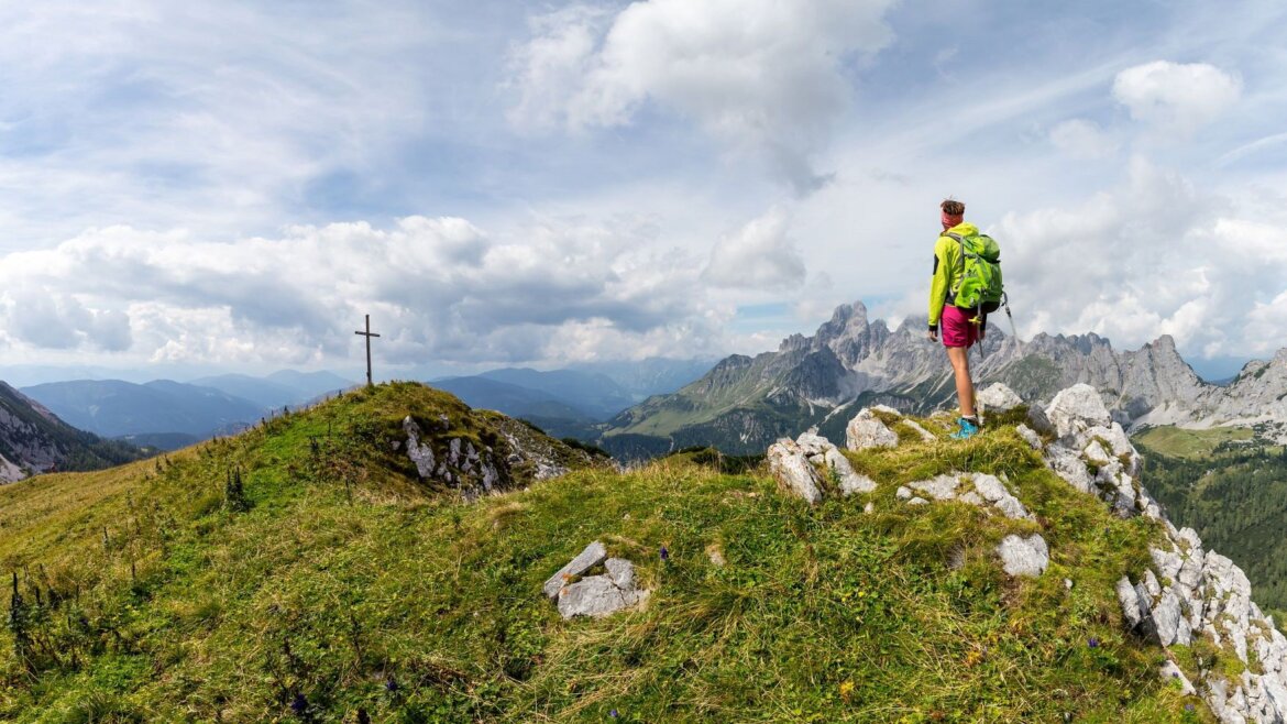 Turista stojí na skalnatém hřebeni v oblasti Filzmoos v Salcbursku a dívá se na vzdálené vrcholy a vrcholový kříž (Zvětšený pohled)