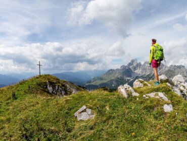 Turista stojí na skalnatém hřebeni v oblasti Filzmoos v Salcbursku a dívá se na vzdálené vrcholy a vrcholový kříž