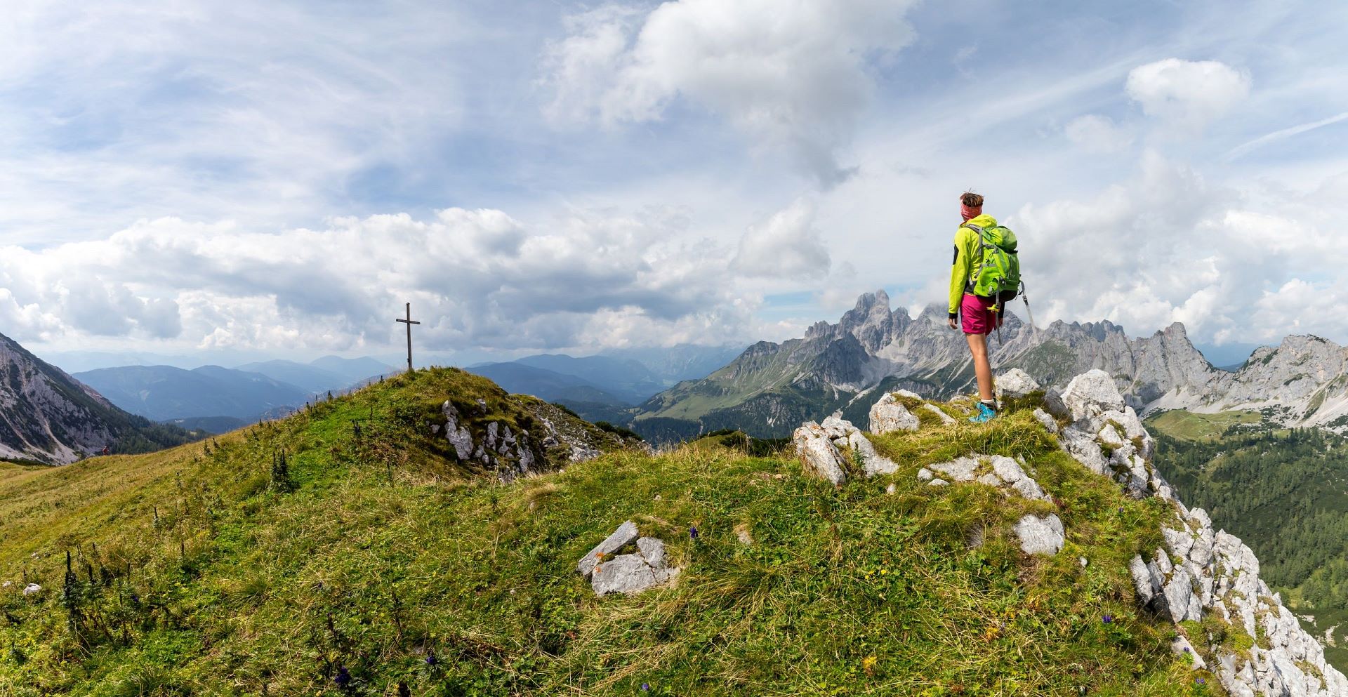 Turista stojí na skalnatém hřebeni v oblasti Filzmoos v Salcbursku a dívá se na vzdálené vrcholy a vrcholový kříž