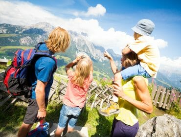 Čtyřčlenná rodina si za slunečného dne užívá malebný výhled na hory, přičemž jedno dítě je na ramenou dospělého.
