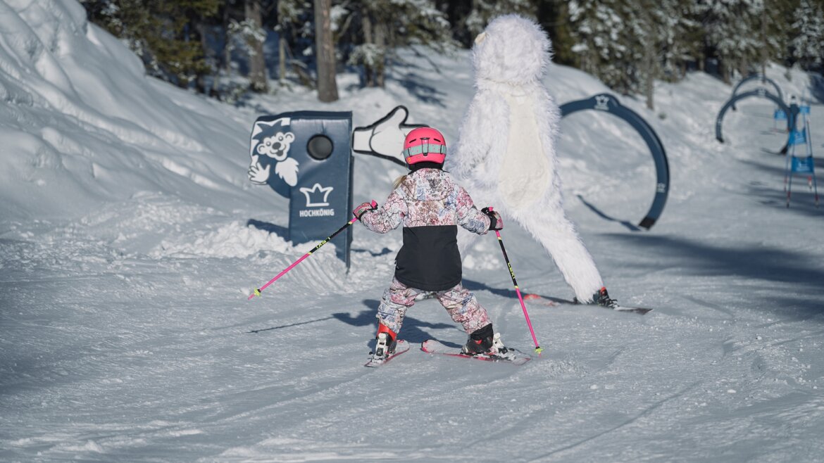 Děti se v Hochkönigu učí první oblouky v hravém a bezpečném prostředí © Hochkönig Tourismus Dítě v růžové přilbě lyžuje vedle maskota yetiho na zasněženém svahu Hochkönigu v zimě. (Zvětšený pohled)