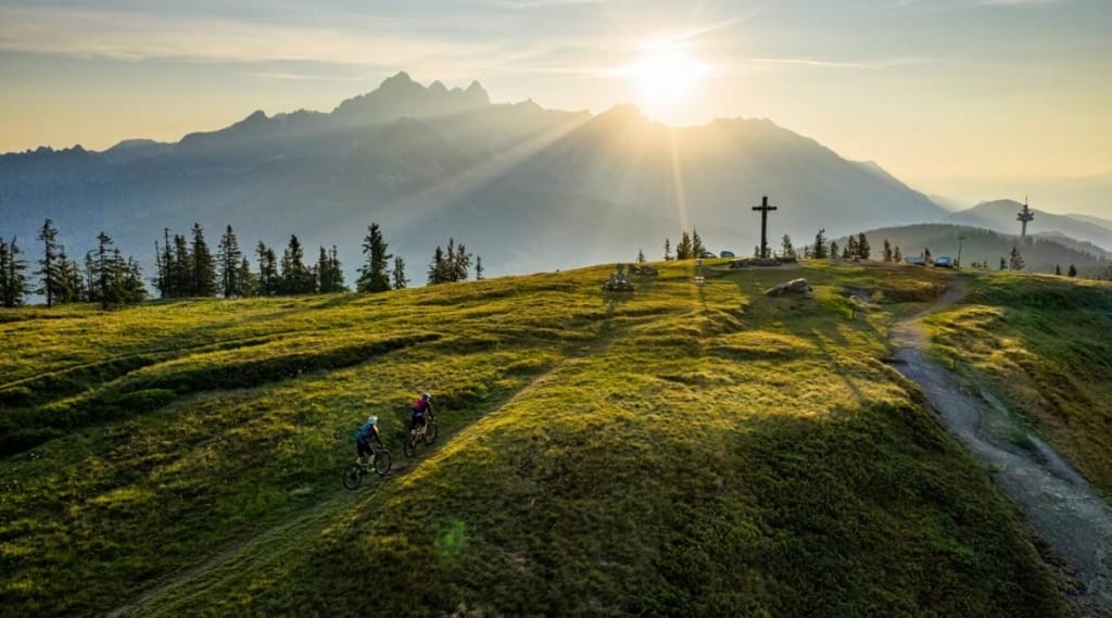 Cyklisté na zelené hoře Rossbrand s vrcholovým křížem a západem slunce 