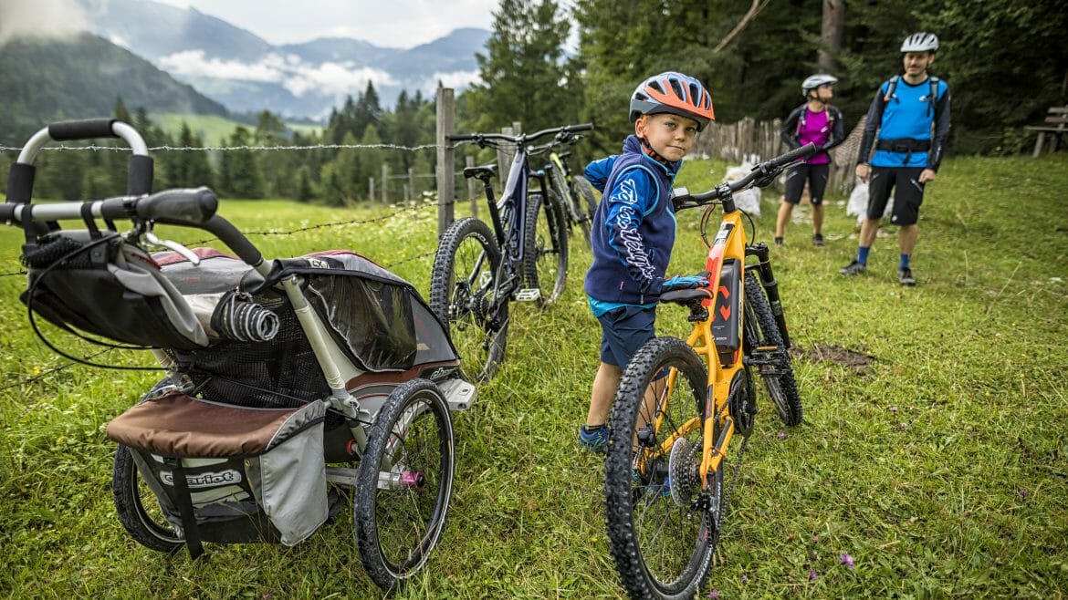 Chlapec s dětským elektrokolem na výletě s rodiči, kteří pro dítě využili i přívěsný vozík za kolo, aby byla cesta do hor co nejpohodlnější 