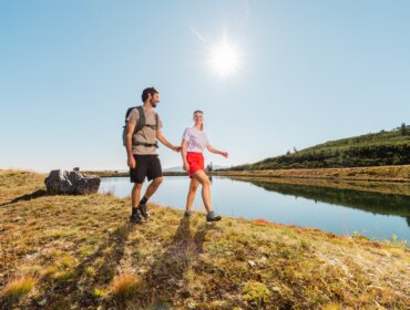 Muž a žena se drží za ruce na túře u jezera v Salcburském Lungau pod jasnou modrou oblohou.